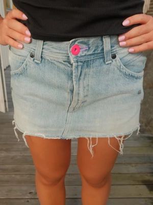 Ljusblå jeanskjol unik - Trendig ljusblå jeanskjol i kort modell med rå, fransig kant nertill. Kjolen har fem fickor, bälteshällor och en rosa knapp framtill som cool detalj. Perfekt för dig som gillar en avslappnad och edgy stil. 