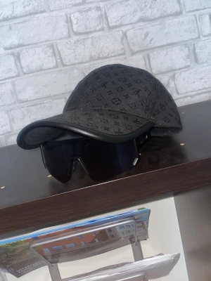 Svart keps med monogrammönster - Snygg svart keps med diskret monogrammönster över hela ytan. Böjd skärm med svart kant och klassisk passform. Perfekt accessoar för en trendig streetwear-look. Materialet ser ut att vara bomull eller en bomullsblandning.