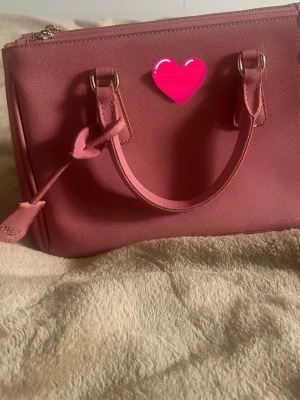 Rosa handväska med gulddetaljer - Snygg rosa handväska i strukturmaterial med två handtag och guldfärgade metalldetaljer. Väskan har dragkedja upptill och en stilren, boxig form. Perfekt för dig som vill ha en trendig accessoar till din outfit.