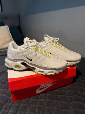 Nike vita Air Max Plus sneakers - Nike Air Max Plus sneakers i vitt mesh med detaljer i grått och neongult. Swoosh-logga på sidan, synliga Air-enheter i sulan och vågiga linjer på ovandelen. Klassisk chunky sula och vita skosnören. Perfekta för dig som gillar streetwear och sportig stil.