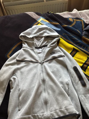 Ljusgrå Nike hoodie med dragkedja - Snygg ljusgrå hoodie från Nike med hel dragkedja framtill och svart Nike-logga på bröstet. Hoodien har huva, fickor på sidorna och en extra ficka med dragkedja på ena ärmen. Tillverkad i mjukt sweatshirtmaterial.