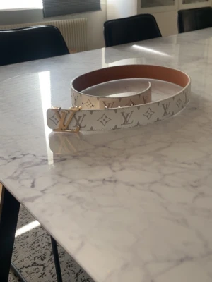 Vitt  bälte med monogram - Säljer ett vitt bälte  med klassiskt monogrammönster och guldfärgat metall spänne. Bältet har en stilren design och är gjort i skinn med beige insida. Perfekt accessoar för att lyfta din outfit.
