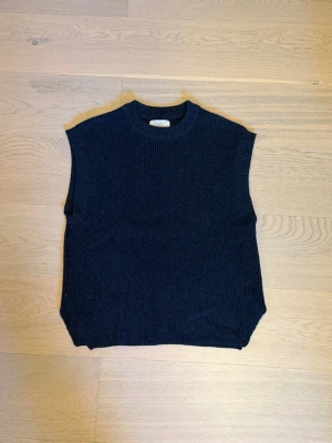 Mörkblå stickad väst - Mörkblå stickad väst med ribbad struktur och rund hals. Västen är ärmlös och har en rak, avslappnad passform. Perfekt att bära över en skjorta eller t-shirt för en trendig lager-på-lager-look.