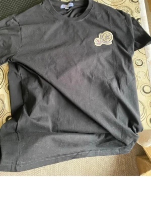 Svart Moncler t-shirt - Svart t-shirt från Moncler. På bröstet sitter Monclers ikoniska logotyp i vitt och beige. Tillverkad i mjukt bomullstyg som känns skönt mot huden. Perfekt för dig som gillar stilrena och exklusiva plagg.