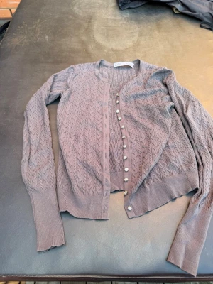 Ljuslila kofta från ZARA - Säljer en ljuslila kofta från ZARA med snyggt stickat mönster och dekorativa knappar framtill. Koftan har långa ärmar och ribbade muddar vid ärmslut och nederkant. Perfekt att slänga över en topp för en chill look.