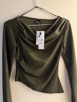 grön långärmad topp från Gina Tricot - Säljer en olivgrön långärmad topp från Gina Tricot med draperad halsringning. Toppen har en figurnära passform och är tillverkad i ett mjukt, stretchigt material. Perfekt för dig som vill ha en stilren och trendig look.(PRIS LAPP KVAR)