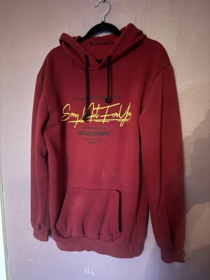 Vinröd hoodie med tryck och huva - Snygg vinröd hoodie med huva och svart dragsko. Framsidan har ett gult och svart tryck med texten 'Sorry Not For You' och 'Establishment'. Stor känguruficka framtill och ribbade muddar. Perfekt för en chill och avslappnad stil. Den har små stains som knappts syns