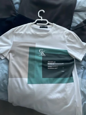 Vit t-shirt Ck - Vit t-shirt med grafiskt tryck i grönt och svart samt textdetaljer på framsidan. Rund halsringning och korta ärmar. Perfekt för en trendig och avslappnad stil.