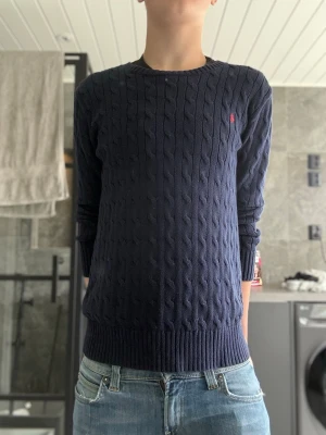 Mörkblå Ralph lauren tröja - Snygg mörkblå kabelstickad tröja med rund halsringning och långa ärmar. Tröjan har ett diskret rött broderat märke på bröstet och ribbade muddar vid ärmslut och nederkant. Perfekt till jeans för en clean och klassisk look.