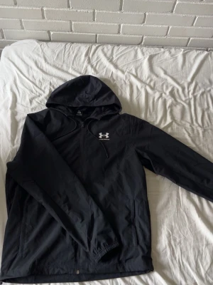 Svart jacka från Under Armour - Säljer en svart hoodie från Under Armour med dragkedja och huva. Hoodien har ett vitt Under Armour-logotyptryck på bröstet och är tillverkad i ett lätt, sportigt material med meshfoder på insidan. Perfekt för träning eller chill.