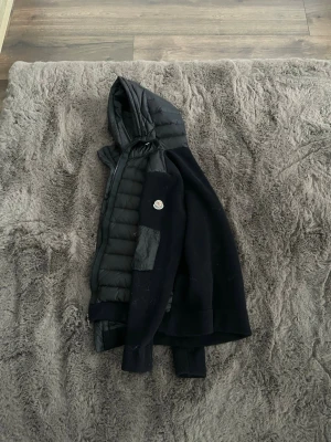 Moncler carding  - Snygg svart dunjacka från Moncler med stickade ärmar och huva. Jackan har quiltad framsida och baksida i dunmaterial, samt ribbade muddar.storlek L passar M Kan tänka mig att gå ner i pris vid en snabb och smidig affär mvh Lucas