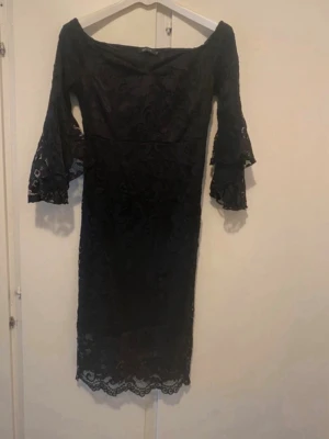 Svart offshoulder spetsklänning - Snygg svart klänning med offshoulder-modell och trekvartsärmar med volang. Klänningen är täckt av spets och har en figurnära passform som ger en elegant siluett. Perfekt för dig som vill sticka ut lite extra.