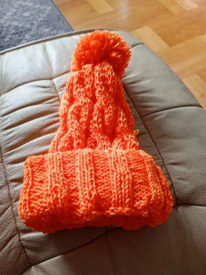 Orange stickad mössa med tofs - Säljer en riktigt snygg orange stickad mössa med stor tofs på toppen. Mössan har ett klassiskt kabelstickat mönster och bred uppvikt kant. Perfekt för dig som vill sticka ut i vinterkylan och hålla dig varm med stil. Material Akrylic och handtvätt 30 grader. Handstickad 