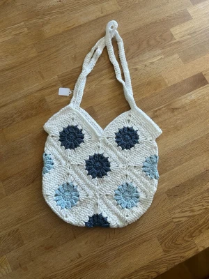 Virkad shoppingväska med blå mönster - Unik handvirkad shoppingväska i vitt med virkade blommor i olika blå nyanser. Väskan har dubbla axelband och en mjuk, rundad form. Perfekt för dig som gillar boho-stil och vill ha något som sticker ut.