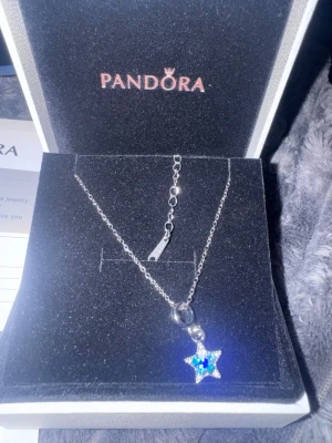 Pandora halsband med blå stjärnhänge - Snyggt halsband från Pandora i silverfärg med ett gnistrande stjärnformat hänge i blått och vitt. Kedjan är tunn och justerbar, och hänget har små stenar som ger extra bling. Perfekt för dig som gillar unika detaljer och vill sticka ut.