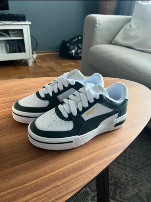 Puma sneakers - CA PRO CLASSIC PUMA SNEAKERS Gröna/vita  Helt oanvända! Nypris 1000. Snygga sneakers från Puma i vitt och mörkgrönt med klassisk siluett. Skorna har perforerad tå, vita skosnören och guldfärgad Puma-logga på sidan. Ovandelen är i läder, och sulan är vit. Perfekta för en sportig och trendig look.