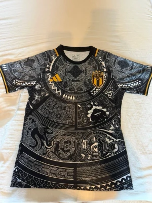 Svart mönstrad Italien-tröja Adidas - Säljer en svart och vit mönstrad fotbollströja från Adidas med Italiens landslagsemblem på bröstet. Tröjan har gula detaljer, rund hals och korta ärmar. Slim fit-modell med unikt grafiskt tryck över hela plagget. Perfekt för dig som vill sticka ut på planen.