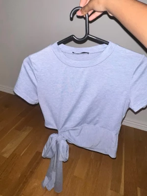 Ljusblå croppad t-shirt med knyt - Säljer en ljusblå croppad t-shirt med korta ärmar och snygg knytning i sidan. Perfekt till jeans eller kjol för en chill och trendig look. Tillverkad i mjuk bomull som känns skön mot huden.