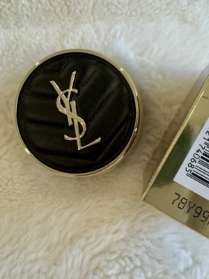 YSL Cushion Foundation svart/guld - Lyxig cushion foundation från Yves Saint Laurent med elegant svart lock i quiltad look och guldiga detaljer. Färg nr #20, Se bild 2 för nyansen. Kompakt rund förpackning med YSL-logga på locket och tillhörande guldig kartong. Medkommer även en sminkpuff med YSL logga (se bild 3). Den är helt ny oöppnad. Perfekt för enkel applicering och touch-ups. 