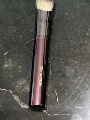 Hourglass vinklad sminkborste - Snygg vinklad sminkborste från Hourglass med mörklila, nästan svart handtag och guldtext. Borsten har täta, mjuka syntetiska strån i beige och vitt, perfekt för contouring eller applicering av foundation. Handtaget är elegant och ergonomiskt utformat.