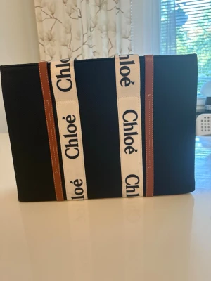  Woody svart handväska - Snygg svart handväska  med vita handtag och logotyp i svart text. Väskan har bruna detaljer i läder och är tillverkad i canvas. Rymlig insida med dragkedjefack och öppen topp. Perfekt för dig som gillar stilrena och trendiga accessoarer.