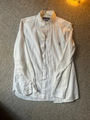 Vit skjorta från Ralph Lauren - Nypris ca 1500kr. Klassisk vit skjorta från Ralph Lauren med button-down krage och broderad marinblå logga på bröstet. Skjortan har långa ärmar, knappar framtill och är tillverkad i bomull. Perfekt för en clean och stilren look.