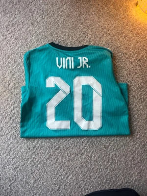 Real Madrid bortatröja Vini Jr #20 -  Nypris Ca 1150kr Snygg turkos Real Madrid bortatröja från Adidas med Vini Jr och nummer 20 på ryggen. Tröjan har vita detaljer, klubbmärke och Adidas-logga på bröstet samt 'Emirates Fly Better' tryckt framtill. Tillverkad i lätt och ventilerande material, perfekt för fotbollsträning.