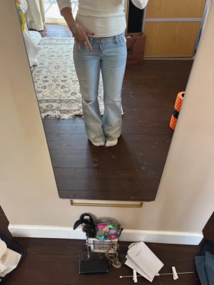 Ljusblå slitna bootcut jeans - Säljer ett par ljusblå bootcut jeans med slitningar på knäna och klassisk femficksmodell. Jeansen har en avslappnad passform och är tillverkade i denim. Perfekta för en trendig och avslappnad stil.