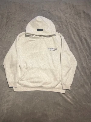 Essentials Fear of God set - Säljer ett snyggt set från Essentials Fear of God i ljusgrå färg. Setet består av en hoodie med känguruficka och tryck på bröstet samt matchande mjukisbyxor med resår i midjan och tryck på benet. Perfekt för en avslappnad och trendig look.