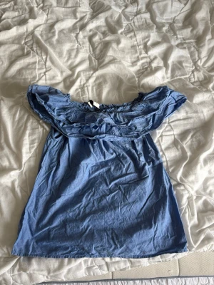 Blå offshoulder topp från Neo Noir - Säljer en blå offshoulder topp från Neo Noir med volangdetaljer längs axlarna. Toppen har en lös passform och är tillverkad i ett mjukt bomullsliknande material. Perfekt för sommarens alla tillfällen!