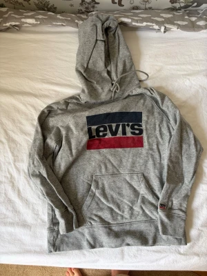 Grå Levi's hoodie med logga - Säljer en grå hoodie från Levi's med stor logga i blått, vitt och rött på bröstet. Hoodien har huva med dragsko och en klassisk magficka framtill. Perfekt för en chill och avslappnad stil.
