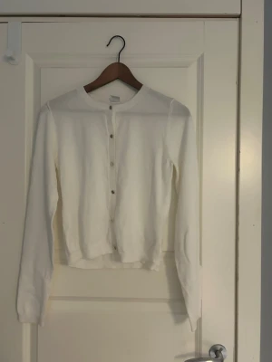 Vit basic kofta med rund hals - Klassisk vit kofta med rund halsringning och knappar framtill. Långärmad modell i mjukt material, perfekt att slänga över en topp eller t-shirt. Enkel och stilren design som passar till det mesta.