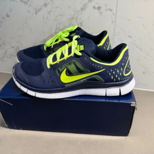 Nike sneakers mörkblå och neon - Snygga sneakers från Nike i mörkblått med neonfärgade detaljer och snören. Skorna har ventilerande meshpartier, vit sula med svart och gulddetaljer undertill samt klassisk Nike-logga på sidan. Perfekta för dig som gillar sportig och modern stil.