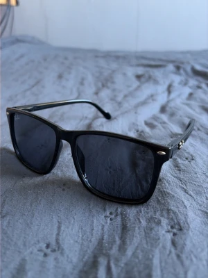 Svarta Ray-Ban solglasögon - Klassiska svarta solglasögon från Ray-Ban med fyrkantig form och mörka glas. Bågarna är i blank svart plast och har små silverdetaljer vid gångjärnen. Perfekta för soliga dagar och ger en tidlös look.