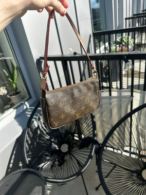 Lv pochete  - Snygg liten handväska från Louis Vuitton i klassiskt brunt monogrammönster. Väskan har en avlång form, guldfärgad dragkedja och ett smalt handtag i brunt och rött läder. Perfekt för dig som vill ha en ikonisk accessoar med lyxig känsla. Är okej skick, bandet börjar spricka lite samt lite brunt inuti men har fof mycket kvar att ge!