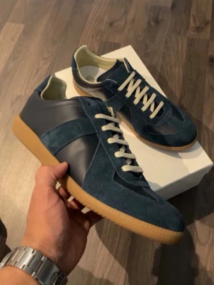 Blå sneakers i mocka och läder - Snygga sneakers i blå mocka och läder med vita snören och klassisk gummisula. Skorna har en låg silhuett och stilren design som passar till många outfits. Insidan är ljus och de har en retrovibe som känns riktigt fräsch. Priset kan diskuteras bara att fråga om funderingar 