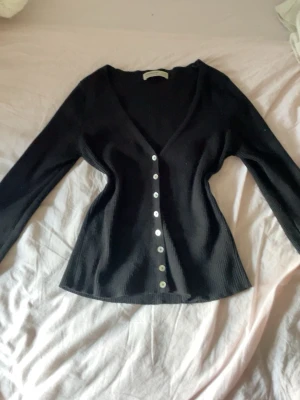 Svart ribbad kofta från Stradivarius - Snygg svart ribbad kofta från Stradivarius med v-ringning och långa ärmar. Koftan har en figurnära passform och knäpps med silvriga knappar framtill. Perfekt att styla över ett linne eller t-shirt för en chill look.