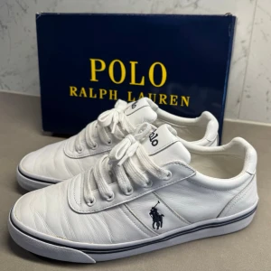 Vita Polo Ralph Lauren sneakers - Stilrena vita sneakers från Polo Ralph Lauren med klassisk låg profil och ikoniska loggan broderad på sidan. Skorna har vita snören, diskreta sömmar och en slitstark gummisula med randig detalj. Perfekta för en clean och fräsch look.