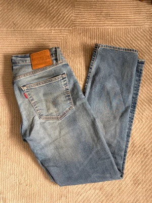 Ljusblå raka jeans från Levi's - Säljer ett par klassiska ljusblå jeans från Levi's med raka ben och femficksmodell. Jeansen har en snygg tvättad look, normal midja och detaljer som den ikoniska röda Levi's-lappen på bakfickan samt läderpatch i midjan. Perfekta för en avslappnad stil.