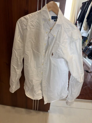 Vit skjorta från Ralph Lauren - Klassisk vit skjorta från Ralph Lauren med button-down krage och broderad logga på bröstet. Tillverkad i bomull och har långa ärmar samt knappar framtill. Perfekt för en clean och stilren look.