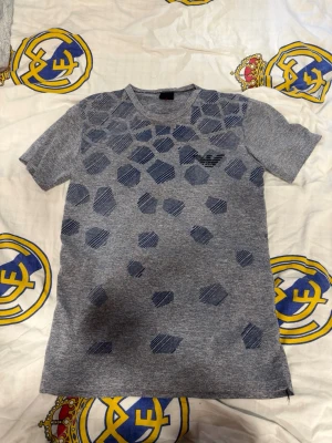Grå armani t-shirt med geometriskt mönster - Snygg grå armani t-shirt med mörkblå geometriska former på framsidan. T-shirten har rund hals och korta ärmar. Tillverkad i mjukt material som känns skönt mot huden. Perfekt för en avslappnad och trendig look.