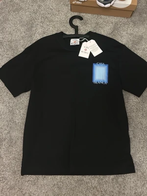 Svart Casa Blanca t-shirt med tryck - Snygg svart t-shirt från Casa Blanca med stort grafiskt tryck i blått och vitt på ryggen samt mindre tryck på bröstet. Klassisk passform och rund hals. Perfekt för dig som gillar stilrena men ändå unika plagg.