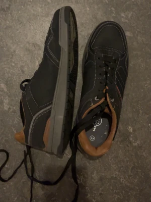 Svarta sneakers med bruna detaljer - Snygga svarta sneakers med bruna detaljer runt hälen och på insidan. Skorna har vita kontrastsömmar och svarta skosnören. Yttermaterialet ser ut att vara en mix av mocka och textil, med en slitstark grå sula. Perfekta för en avslappnad streetstil.