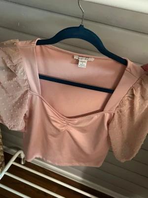Ljusrosa topp med puffärmar - Supersöt ljusrosa topp med transparenta puffärmar i mesh och små prickar. Toppen har en v-ringad urringning med rynkdetalj framtill och är croppad i modellen. Perfekt för dig som vill ha en feminin och trendig look.