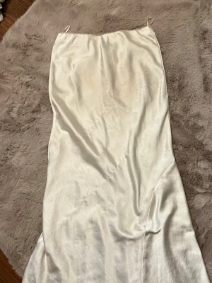 Vit långklänning i satin - Elegant vit långklänning med tunna axelband och slät, glansig yta i satinliknande material. Klänningen har en enkel och stilren design som ger en lyxig känsla. Perfekt för dig som vill ha en minimalistisk look.