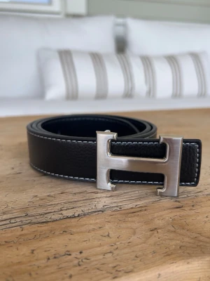 Svart Herme’s skärp med silverfärgat H-spänne - Snyggt svart Herme’s bälte i skinn med ett stort, silverfärgat H-format spänne. Bältet har vita kontrastsömmar längs kanterna som ger en stilren look. Perfekt accessoar för att lyfta vilken outfit som helst.