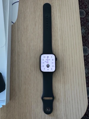 Apple Watch 7 45mm GPS - Stilren svart smartklocka med rektangulär skärm och svart silikonarmband. Klockan har en digital urtavla med tydliga siffror och en knapp på sidan. Perfekt för dig som vill ha koll på tiden och notiser direkt på handleden.