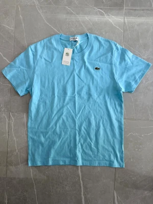 Ljusblå t-shirt från Lacoste - Snygg ljusblå t-shirt från Lacoste med klassisk krokodillogga på bröstet. Tillverkad i mjuk bomull och har en normal passform med korta ärmar och rund hals. Perfekt för en clean och fräsch look.
