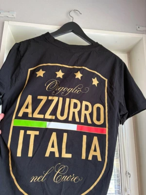 Svart t-shirt med Italiendesign - Svart t-shirt med stort tryck på ryggen och mindre tryck på bröstet. Texten 'Azzurro Italia' och italienska flaggans färger syns tydligt. Rund hals och korta ärmar. Perfekt för dig som vill visa kärlek till Italien.