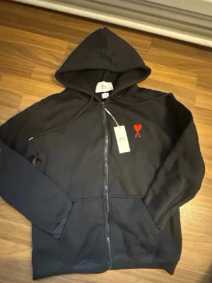 Ami - zip up hoodie - Snygg svart hoodie från Ami Alexandre Mattiussi med dragkedja framtill och huva med dragsko. På bröstet finns Ami:s ikoniska röda hjärta med bokstaven A. Tillverkad i mjuk bomullsblandning och har två fickor framtill.